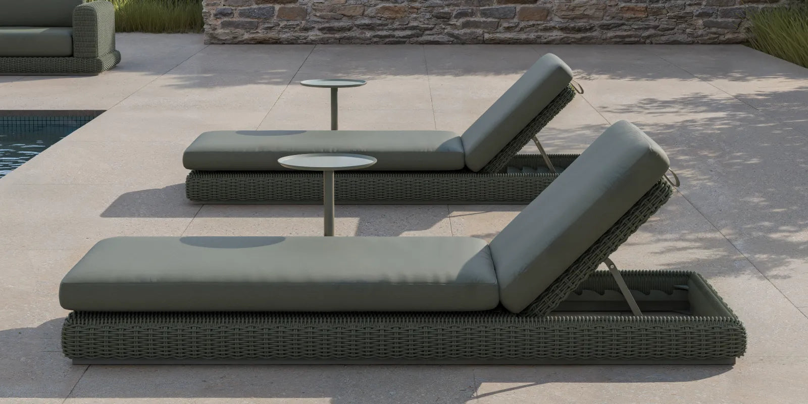 Wide poolside banner of two Neighbor Roundabout Sun Loungers in dark olive-gray resin wicker with matching olive-gray cushions arranged side-by-side on a sunlit stone-tile pool deck, with a small white round side table between them, ornamental grasses to the right, and a stacked-stone wall in the background.