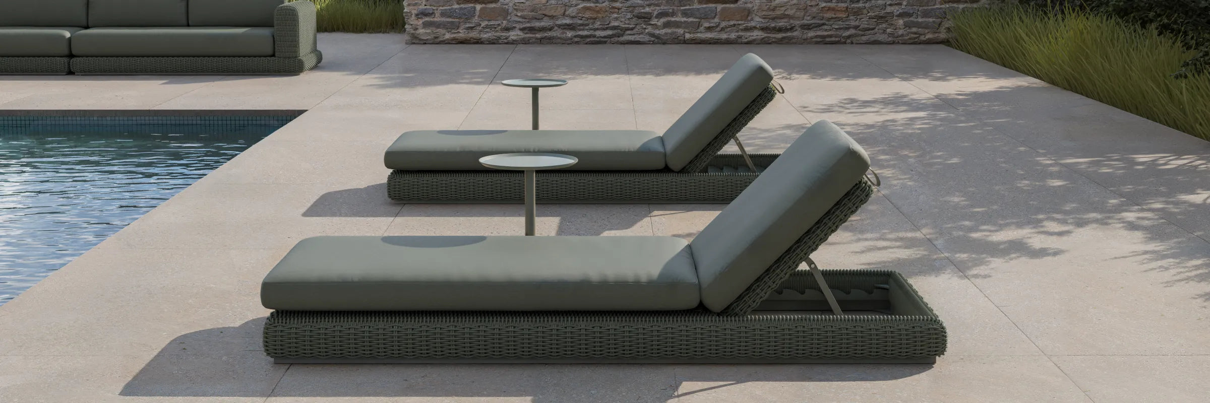Wide poolside banner of two Neighbor Roundabout Sun Loungers in dark olive-gray resin wicker with matching olive-gray cushions arranged side-by-side on a sunlit stone-tile pool deck, with a small white round side table between them, ornamental grasses to the right, and a stacked-stone wall in the background.