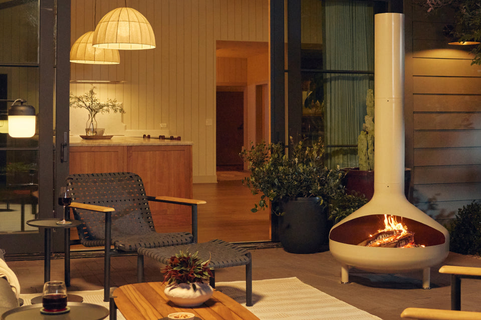 Wide evening patio banner showing a cream-beige Neighbor Totem Chiminea with fire glowing in its firebox on a covered terrace, with woven-strap Hopper Chairs and a wood-and-steel Hopper Coffee Table on a striped outdoor rug to the left and a stacked-stone wall and pendant lights behind.