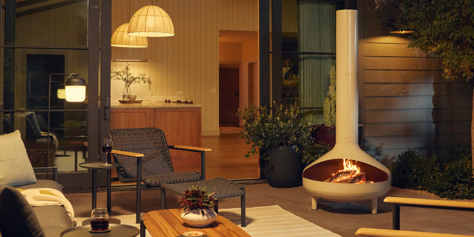 Wide evening patio banner showing a cream-beige Neighbor Totem Chiminea with fire glowing in its firebox on a covered terrace, with woven-strap Hopper Chairs and a wood-and-steel Hopper Coffee Table on a striped outdoor rug to the left and a stacked-stone wall and pendant lights behind.