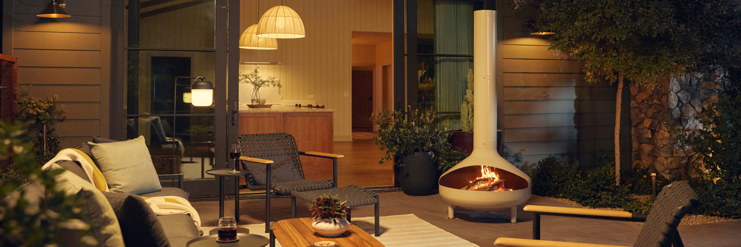 Wide evening patio banner showing a cream-beige Neighbor Totem Chiminea with fire glowing in its firebox on a covered terrace, with woven-strap Hopper Chairs and a wood-and-steel Hopper Coffee Table on a striped outdoor rug to the left and a stacked-stone wall and pendant lights behind.