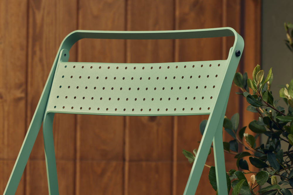 Tight detail of the perforated sage-green steel backrest of a Neighbor Take Two Bistro Chair against a stained-wood plank wall, with glossy laurel foliage just out of focus at right.