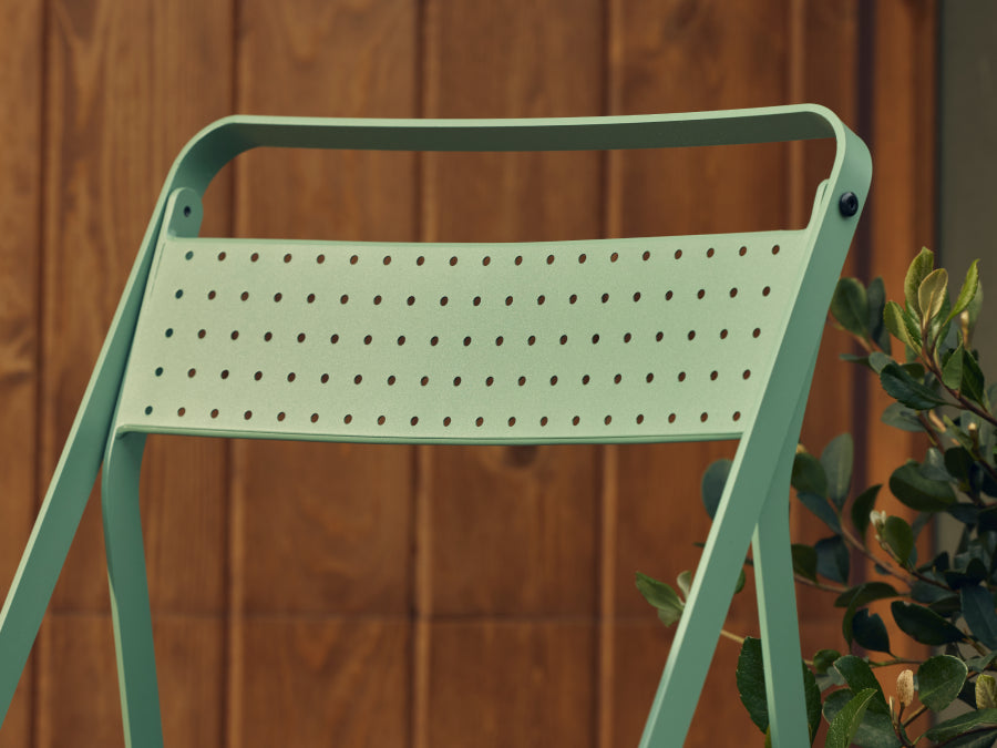 Tight detail of the perforated sage-green steel backrest of a Neighbor Take Two Bistro Chair against a stained-wood plank wall, with glossy laurel foliage just out of focus at right.