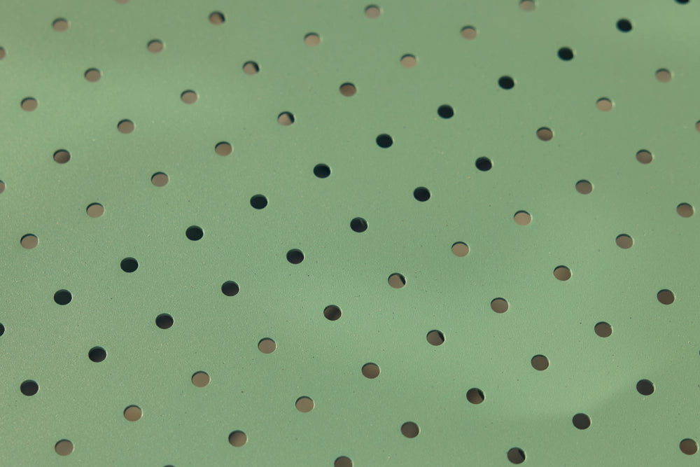 Macro detail of the perforated sage-green powder-coated steel surface of a Neighbor Take Two Bistro Table, raked sunlight glancing across the round laser-cut holes.