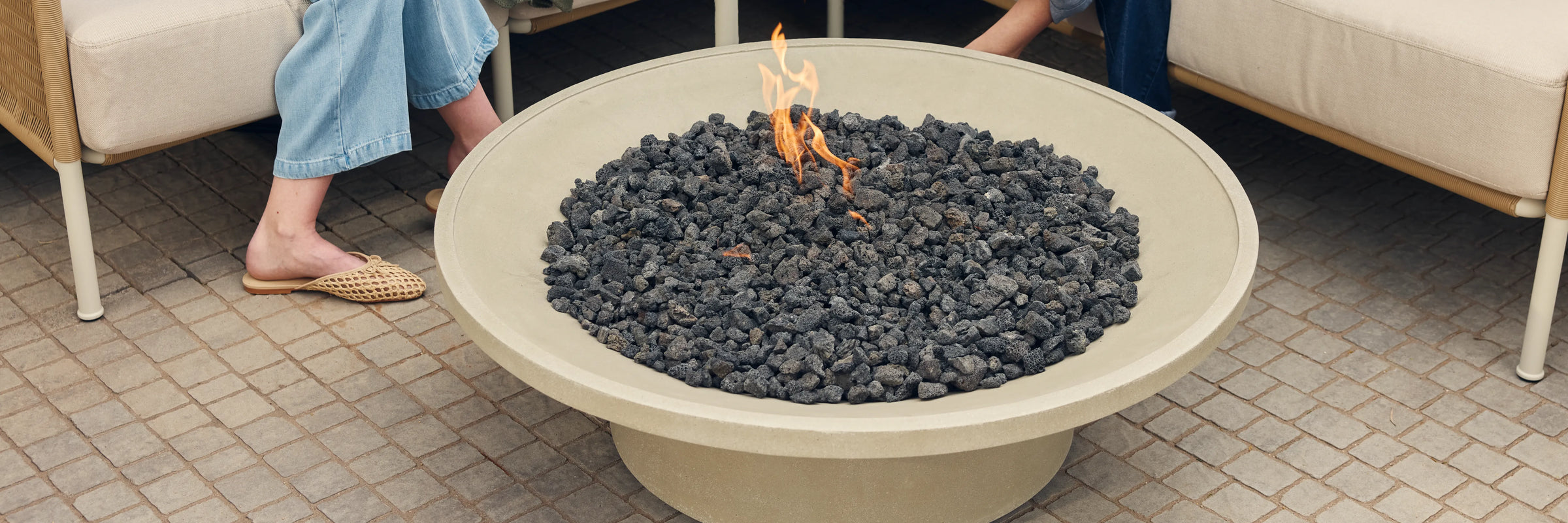 Closeup of Neighbor Brimfire table filled with lava rocks and open flame on a cobblestone patio, Neighbor Camber Sectional visible at edge