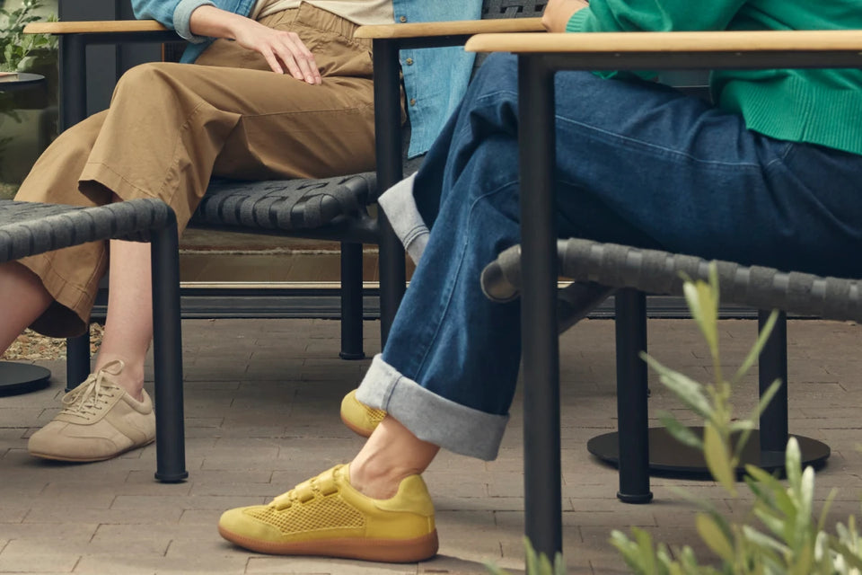 Wide cropped banner showing the lower halves of two friends seated on Neighbor Up-Down Chairs with charcoal woven-strap seats, black powder-coated tubular frames, and natural-wood armrests on a brick-paver patio, one with feet propped on a Neighbor Up-Down Ottoman with matching charcoal woven-strap top, with olive shrubs, a small black side table, and the edge of a cream-beige Totem Chiminea in frame.