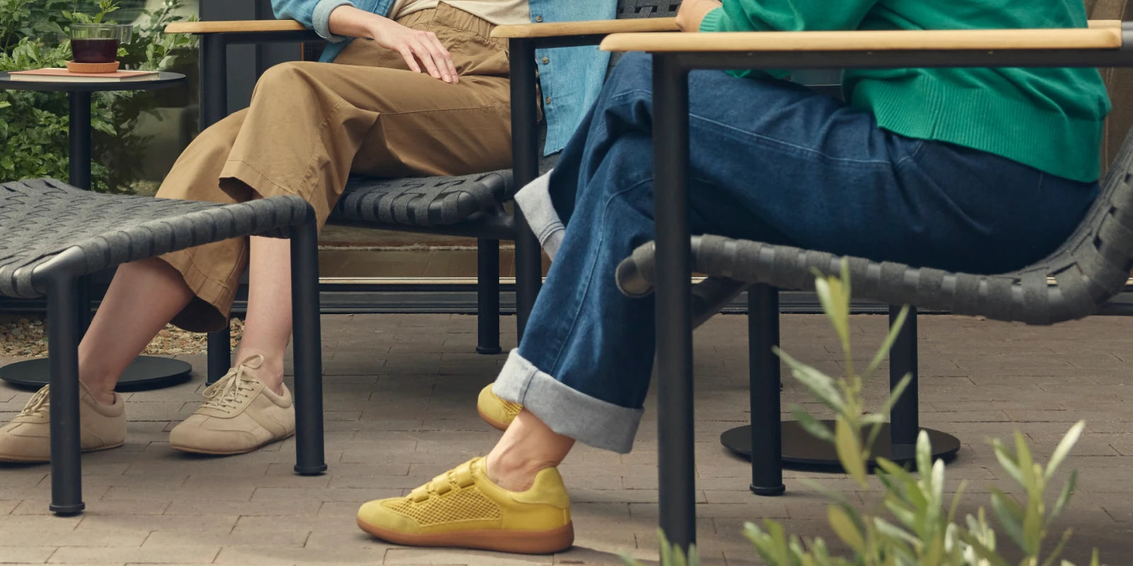 Wide cropped banner showing the lower halves of two friends seated on Neighbor Up-Down Chairs with charcoal woven-strap seats, black powder-coated tubular frames, and natural-wood armrests on a brick-paver patio, one with feet propped on a Neighbor Up-Down Ottoman with matching charcoal woven-strap top, with olive shrubs, a small black side table, and the edge of a cream-beige Totem Chiminea in frame.