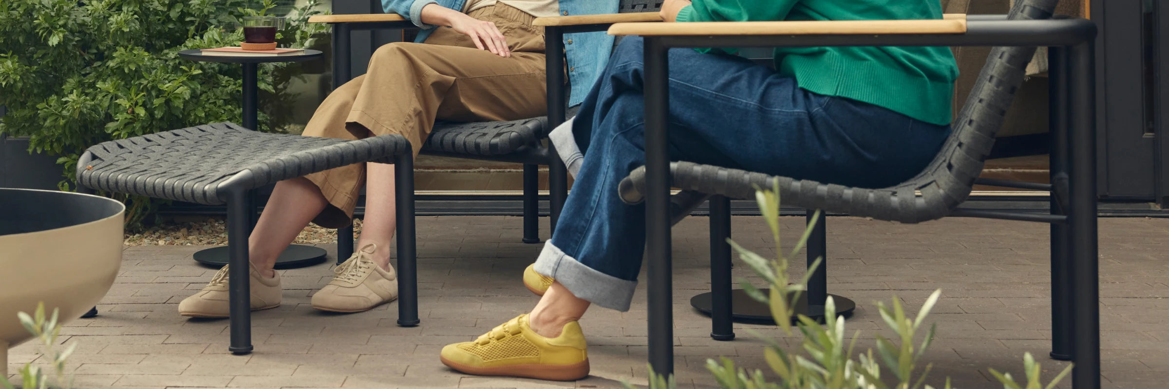 Wide cropped banner showing the lower halves of two friends seated on Neighbor Up-Down Chairs with charcoal woven-strap seats, black powder-coated tubular frames, and natural-wood armrests on a brick-paver patio, one with feet propped on a Neighbor Up-Down Ottoman with matching charcoal woven-strap top, with olive shrubs, a small black side table, and the edge of a cream-beige Totem Chiminea in frame.