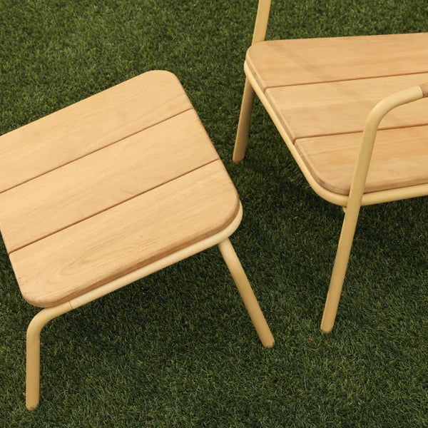 Close detail shot of a Neighbor Hopper Ottoman with natural-wood slat top and yellow powder-coated tubular steel frame placed on artificial-turf grass beside the matching Neighbor Hopper Chair, showing the seamless visual pairing of the two pieces.