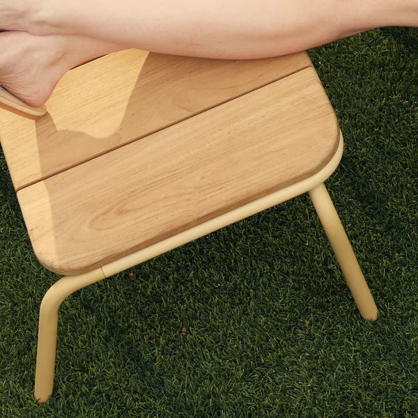 Overhead detail of a woman's bare feet resting on a Neighbor Hopper Ottoman with natural-wood slat top and yellow powder-coated tubular steel frame on artificial-turf grass, capturing the relaxed feel of the wood underfoot.