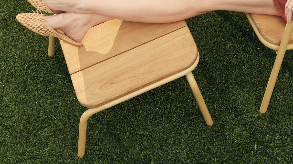 Overhead detail of a woman's bare feet resting on a Neighbor Hopper Ottoman with natural-wood slat top and yellow powder-coated tubular steel frame on artificial-turf grass, capturing the relaxed feel of the wood underfoot.