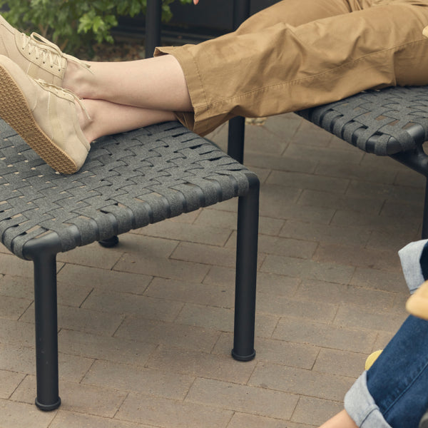 Detail of a Neighbor Up-Down Ottoman in charcoal woven-strap with black powder-coated tubular steel frame on a brick-paver patio, with a woman's outstretched legs and tan sneakers resting on top, paired with the matching Up-Down Chair just behind.