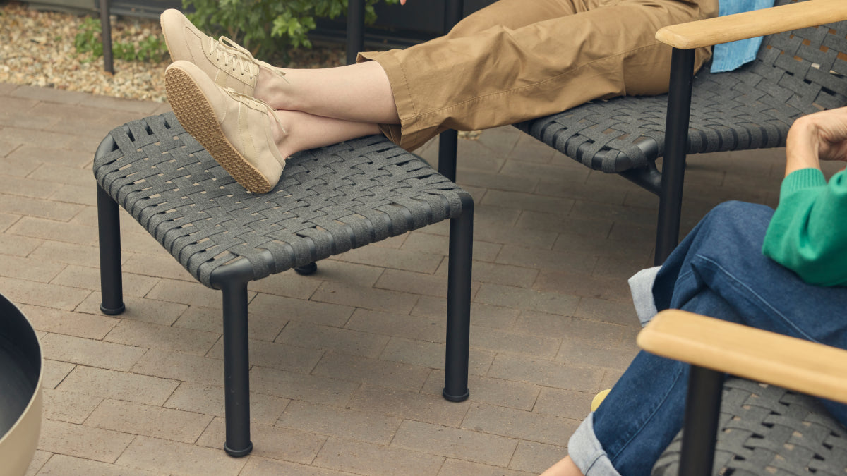 Detail of a Neighbor Up-Down Ottoman in charcoal woven-strap with black powder-coated tubular steel frame on a brick-paver patio, with a woman's outstretched legs and tan sneakers resting on top, paired with the matching Up-Down Chair just behind.