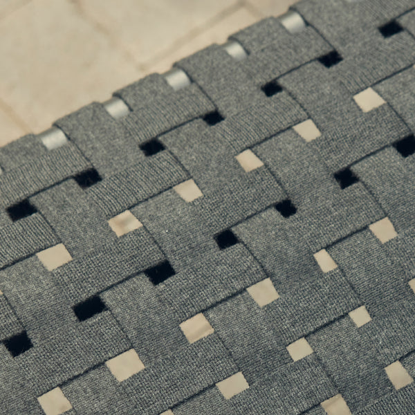 Macro detail of the Neighbor Up-Down Ottoman's charcoal woven-strap top, showing the wide flat straps interlaced in a herringbone pattern over a stone-tile patio surface visible through the openings.