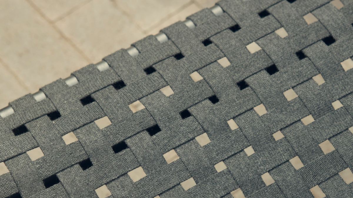 Macro detail of the Neighbor Up-Down Ottoman's charcoal woven-strap top, showing the wide flat straps interlaced in a herringbone pattern over a stone-tile patio surface visible through the openings.