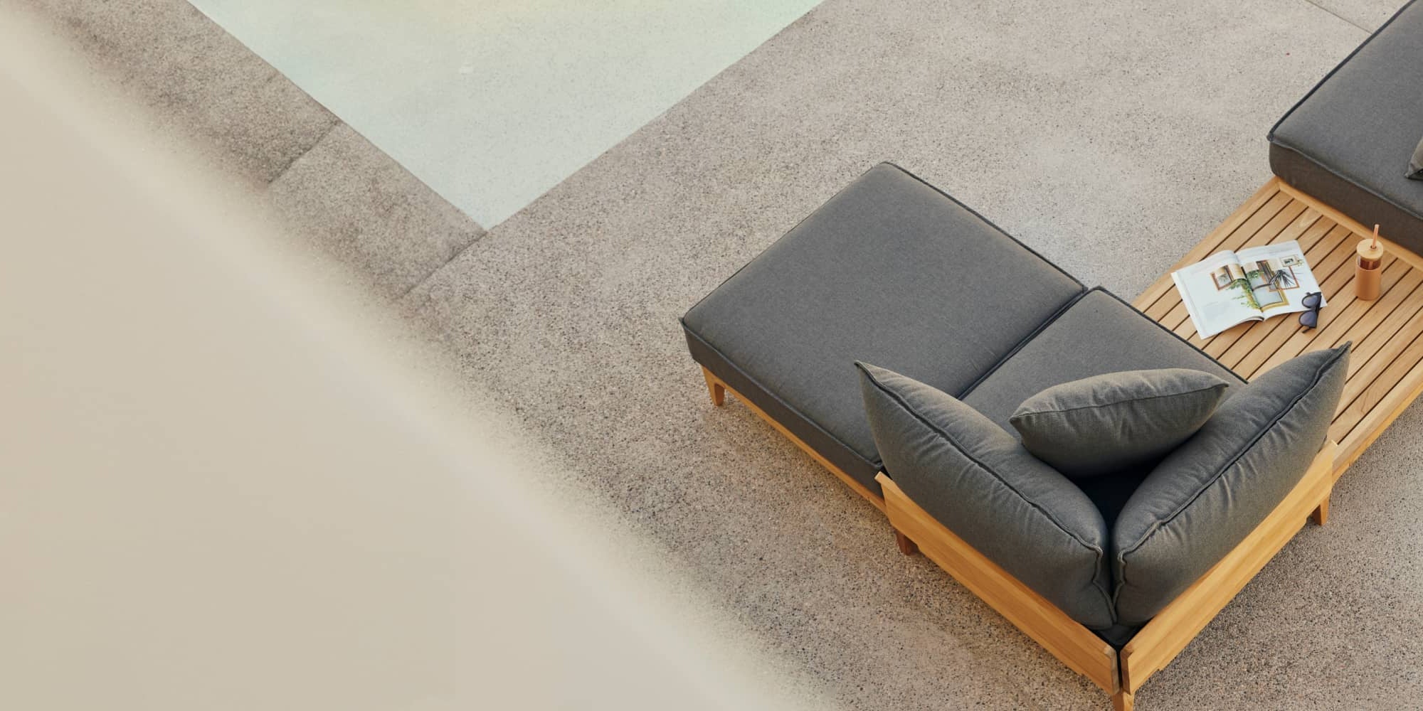 Teak outdoor chaise lounge chair with billowy gray cushions alongside a pool on a patio, photographed from above.