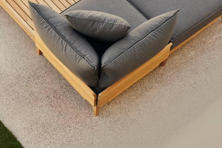 Teak outdoor chaise lounge chair with billowy gray cushions alongside a pool on a patio, photographed from above.