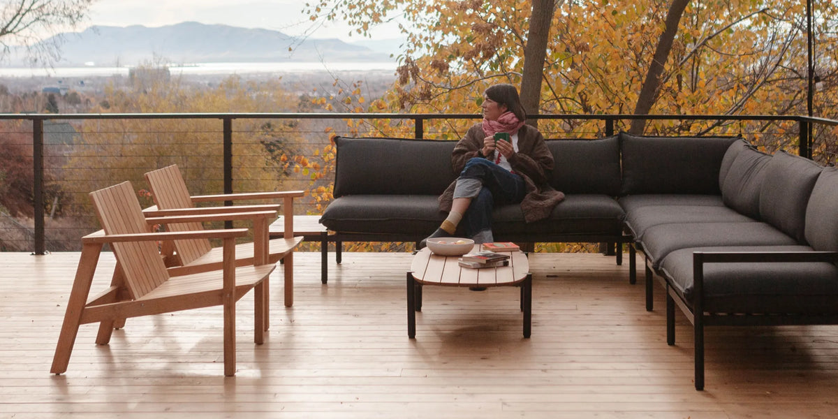 Outdoor patio furniture set on a wooden deck with Amanda Jane Jones sitting on a dark gray sectional sofa, surrounded by autumn trees and a scenic view in the background.