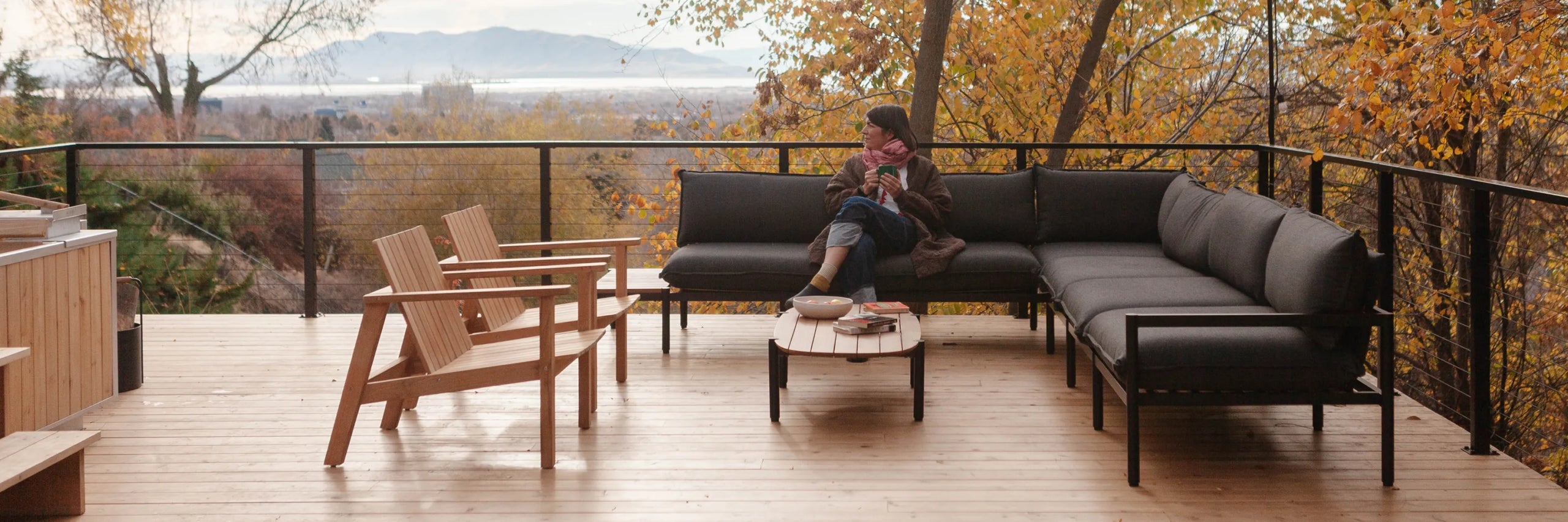 Outdoor patio furniture set on a wooden deck with Amanda Jane Jones sitting on a dark gray sectional sofa, surrounded by autumn trees and a scenic view in the background.