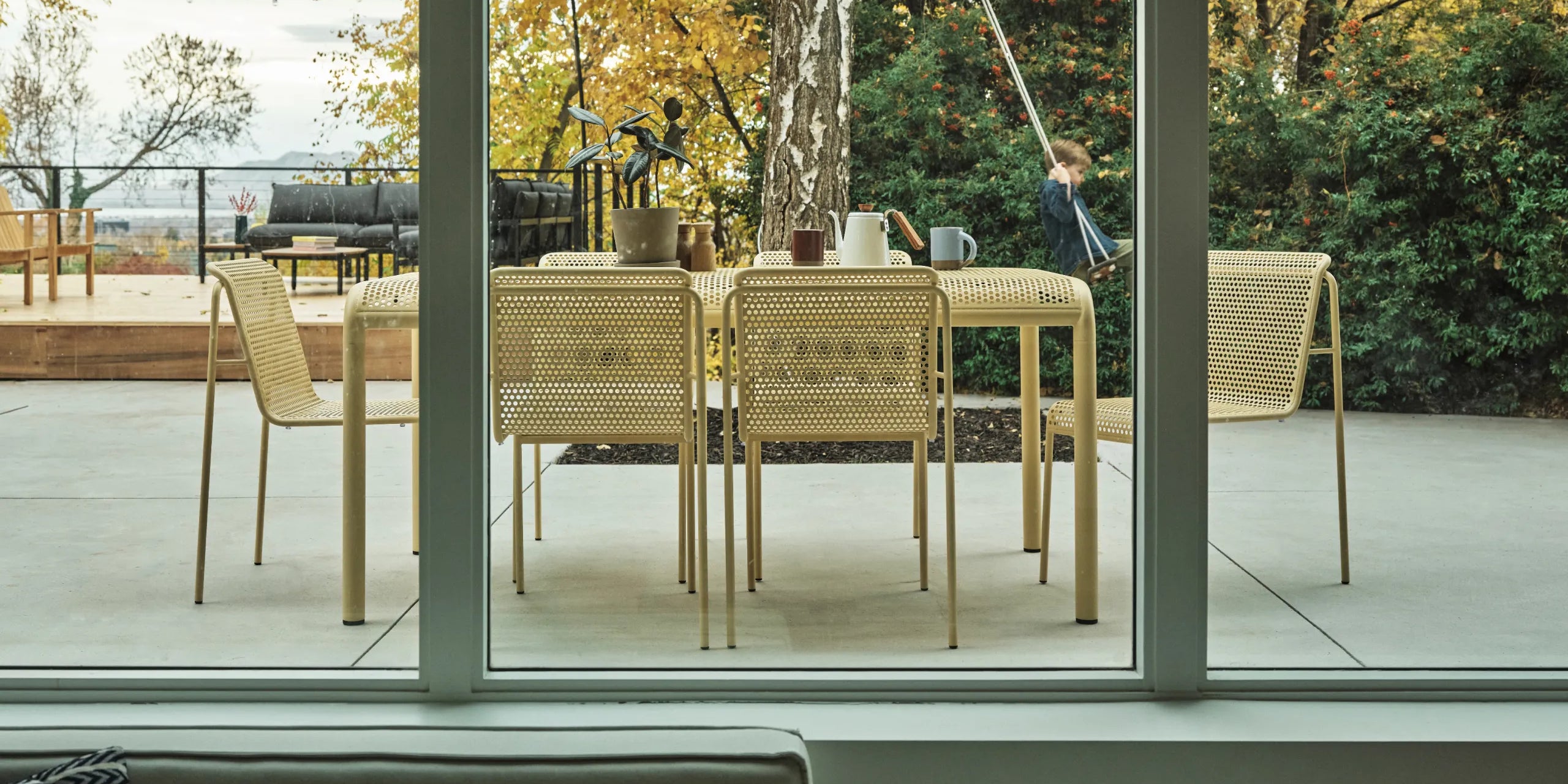 A modern outdoor dining set consisting of a rectangular perforated table and six perforated chairs, shown on a patio with a child sitting on a swing in the background.