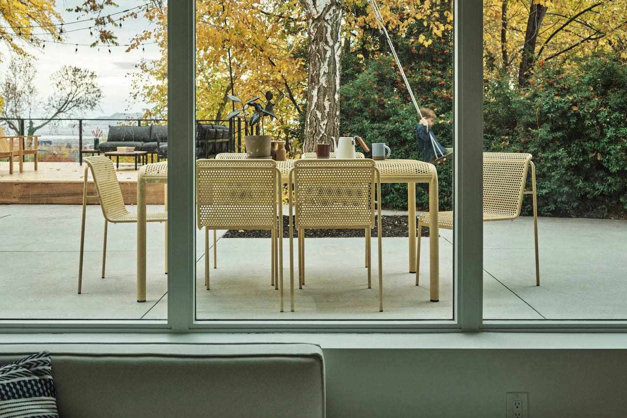 A modern outdoor dining set consisting of a rectangular perforated table and six perforated chairs, shown on a patio with a child sitting on a swing in the background.