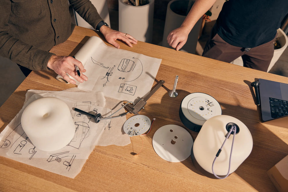 Hands sketching product designs with compass and circular templates on wooden workstation with materials