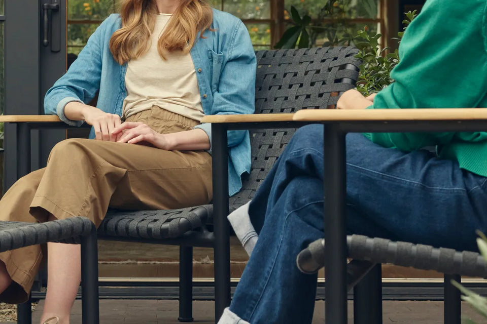 Wide showcase banner of two friends in conversation on a covered patio, seated in Neighbor Up-Down Chairs with charcoal woven-strap seats and natural-wood armrests on black powder-coated tubular frames, with a Neighbor Up-Down Ottoman between them, a small black side table holding a glass and book, and the edge of a cream-beige Totem Chiminea, against a glass-walled garden building backdrop.