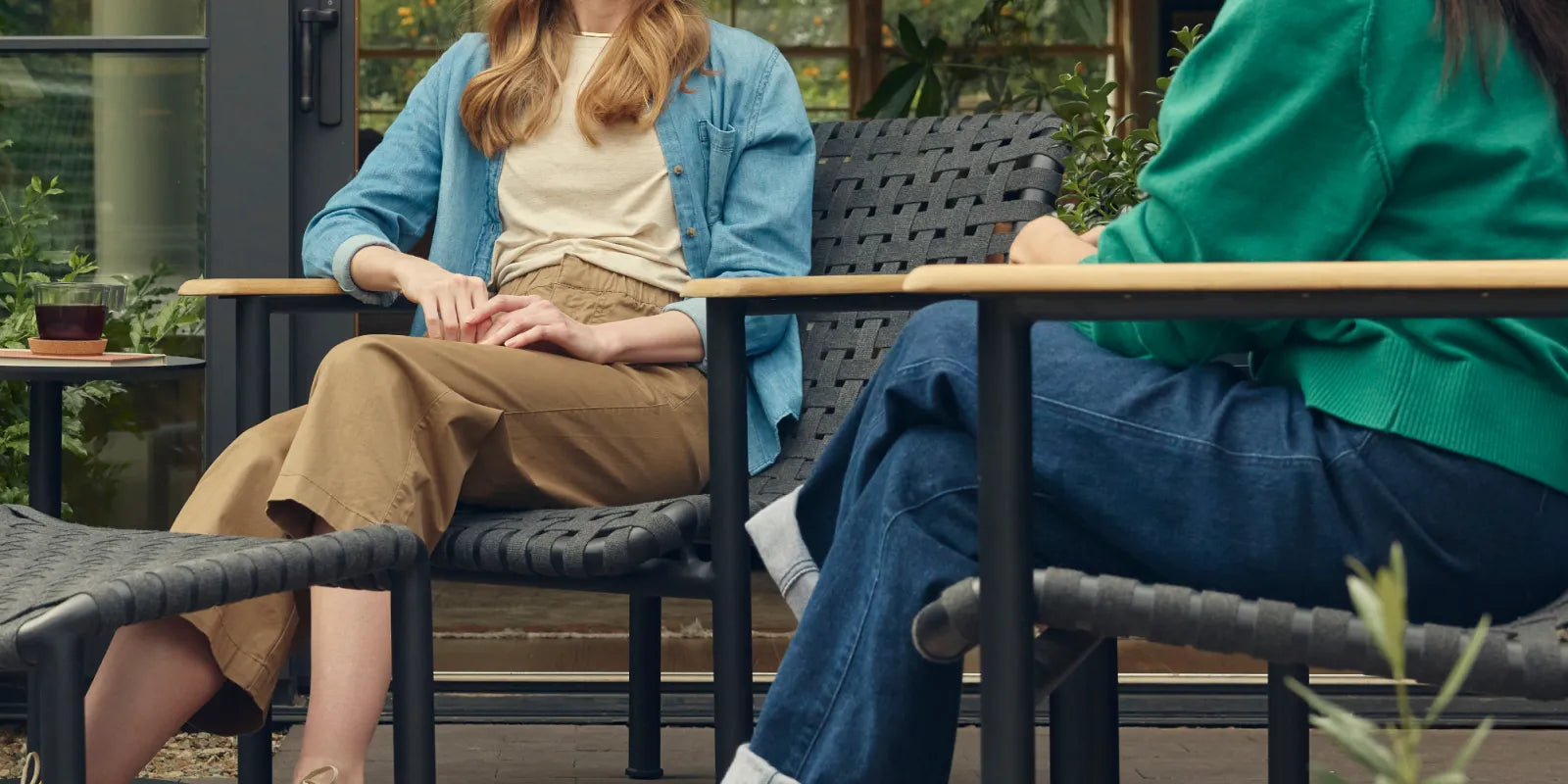 Wide showcase banner of two friends in conversation on a covered patio, seated in Neighbor Up-Down Chairs with charcoal woven-strap seats and natural-wood armrests on black powder-coated tubular frames, with a Neighbor Up-Down Ottoman between them, a small black side table holding a glass and book, and the edge of a cream-beige Totem Chiminea, against a glass-walled garden building backdrop.