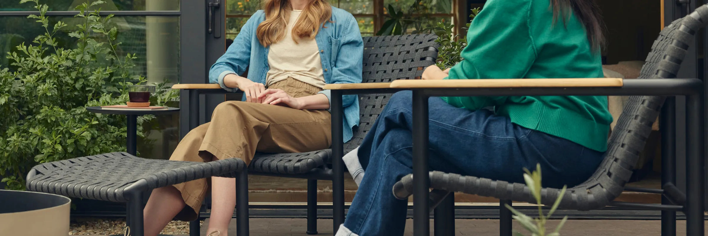 Wide showcase banner of two friends in conversation on a covered patio, seated in Neighbor Up-Down Chairs with charcoal woven-strap seats and natural-wood armrests on black powder-coated tubular frames, with a Neighbor Up-Down Ottoman between them, a small black side table holding a glass and book, and the edge of a cream-beige Totem Chiminea, against a glass-walled garden building backdrop.