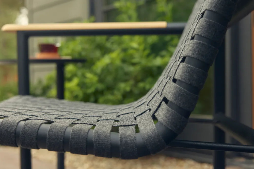 Side close-up of a Neighbor Up-Down Chair with charcoal woven-strap seat and back on a black powder-coated tubular frame, focused on the curved sling profile, with a soft-focus garden patio scene of green shrubs and a black side table in the background.