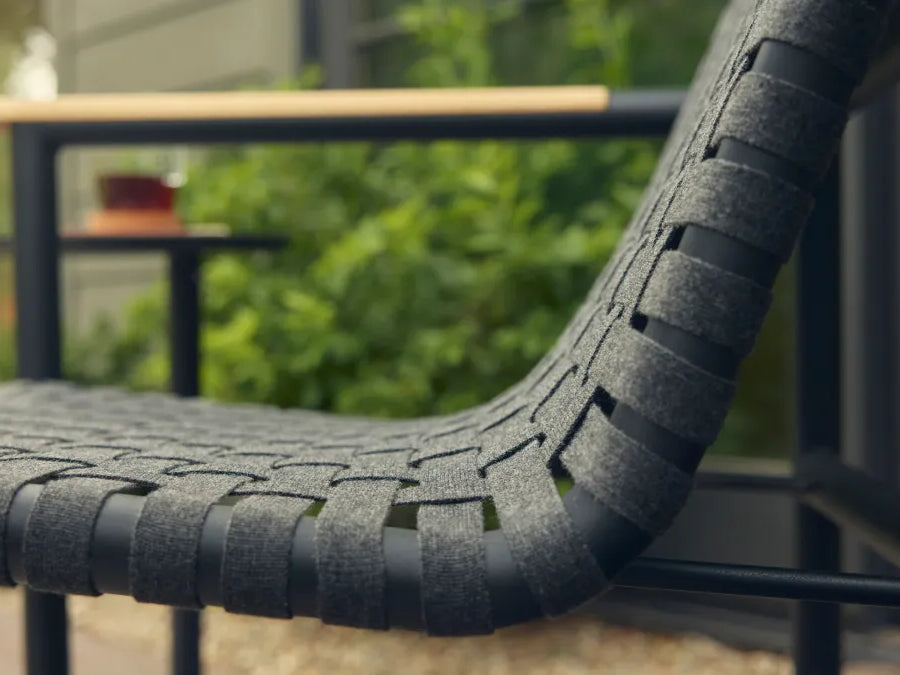 Side close-up of a Neighbor Up-Down Chair with charcoal woven-strap seat and back on a black powder-coated tubular frame, focused on the curved sling profile, with a soft-focus garden patio scene of green shrubs and a black side table in the background.