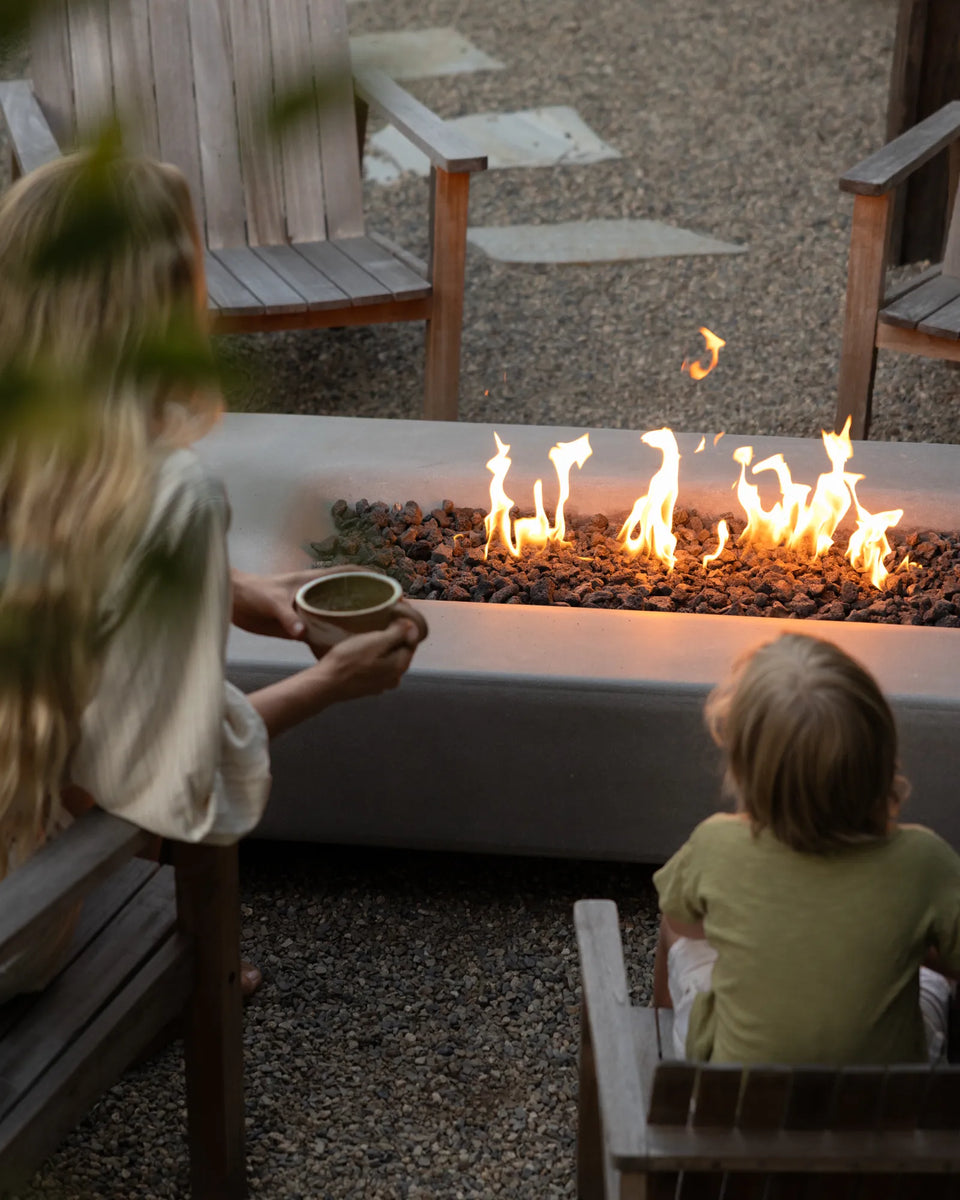 Taylr Anne Castro and her child sitting on a patio beside a modern rectangular fire pit with flames burning, surrounded by wooden chairs and gravel.