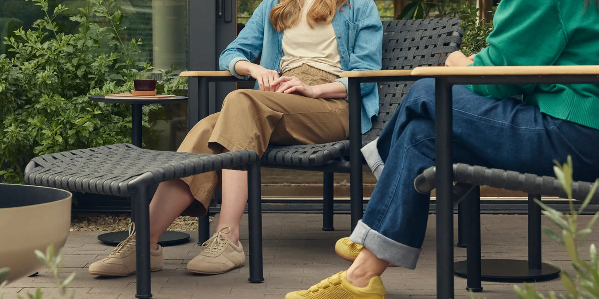 Wide cropped hero of two friends seated and in conversation on Neighbor Up-Down Chairs with charcoal woven-strap seats and natural-wood armrests on black powder-coated tubular frames, with a Neighbor Up-Down Ottoman and small black side table on a brick-paver patio, the edge of a cream-beige Totem Chiminea visible at left.