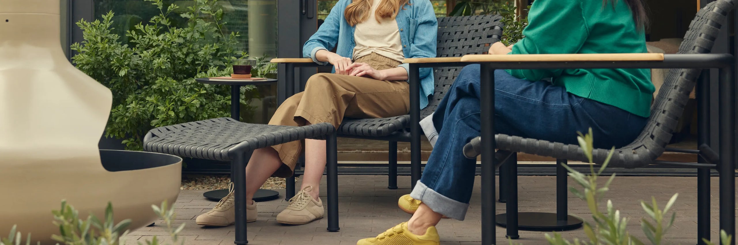 Wide cropped hero of two friends seated and in conversation on Neighbor Up-Down Chairs with charcoal woven-strap seats and natural-wood armrests on black powder-coated tubular frames, with a Neighbor Up-Down Ottoman and small black side table on a brick-paver patio, the edge of a cream-beige Totem Chiminea visible at left.