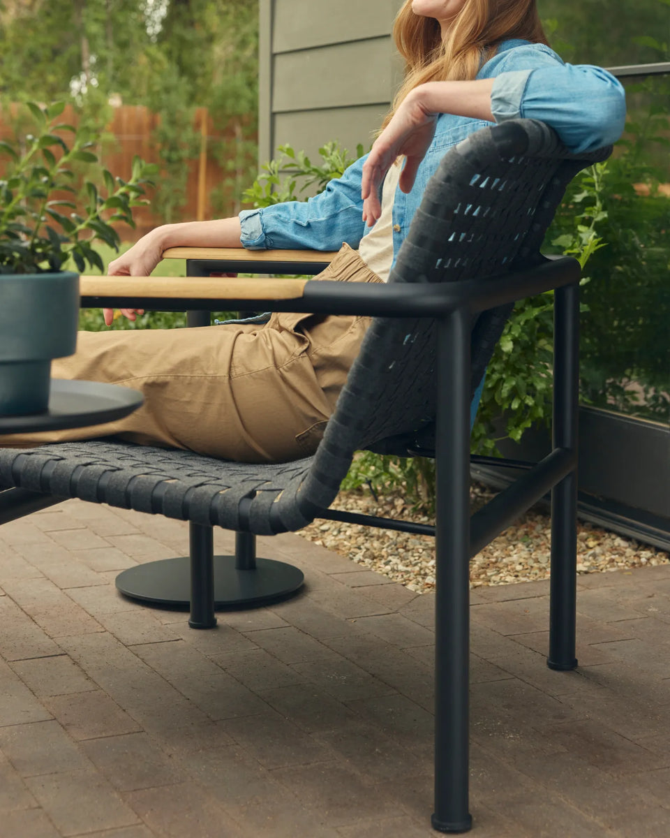 Vertical hero of a woman in a chambray shirt and tan pants reclining in a Neighbor Up-Down Chair with charcoal woven-strap seat-and-back, natural-wood armrest, and black powder-coated tubular steel frame, paired with a Neighbor Up-Down Ottoman and small black side table holding a planter on a brick-paver patio, with green shrubs and a wood-clad home exterior in the background.