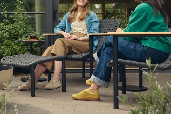 Hero scene of two friends laughing together on Neighbor Up-Down Chairs with charcoal woven-strap seats and natural-wood armrests on black powder-coated tubular frames, a Neighbor Up-Down Ottoman between them and a small black side table holding a glass of iced tea, on a brick-paver patio outside a glass-walled garden building, with the edge of a cream-beige Totem Chiminea at left.