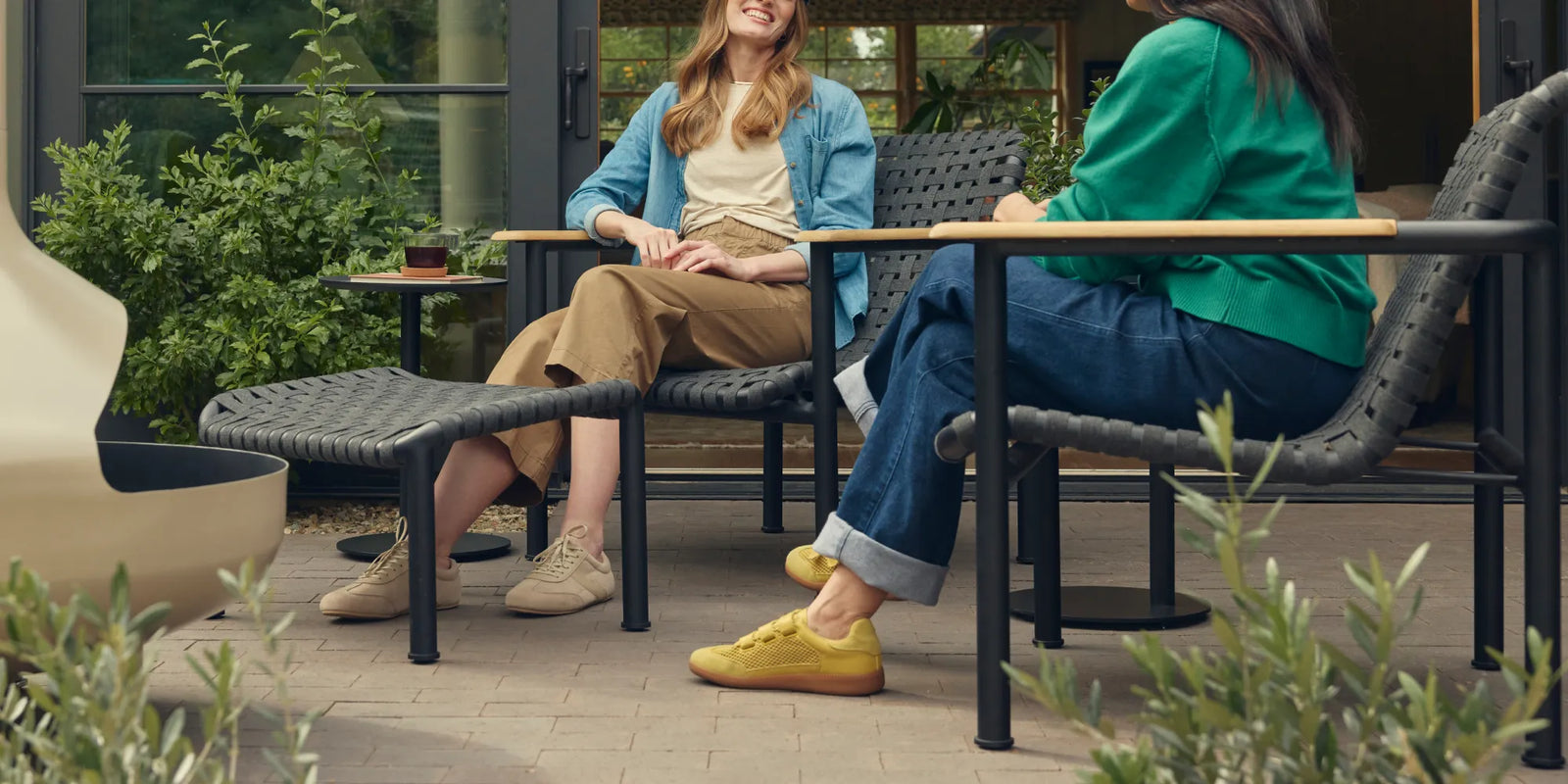 Hero scene of two friends laughing together on Neighbor Up-Down Chairs with charcoal woven-strap seats and natural-wood armrests on black powder-coated tubular frames, a Neighbor Up-Down Ottoman between them and a small black side table holding a glass of iced tea, on a brick-paver patio outside a glass-walled garden building, with the edge of a cream-beige Totem Chiminea at left.