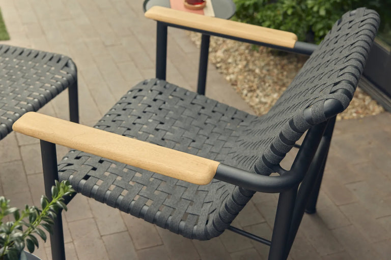 Overhead three-quarter view of a Neighbor Up-Down Chair with charcoal woven-strap seat-and-back sling and natural-wood armrests on a black powder-coated tubular frame, paired with a Neighbor Up-Down Ottoman, both on a brick-paver patio with greenery and a small black side table holding a glass and a coral-orange book in frame.
