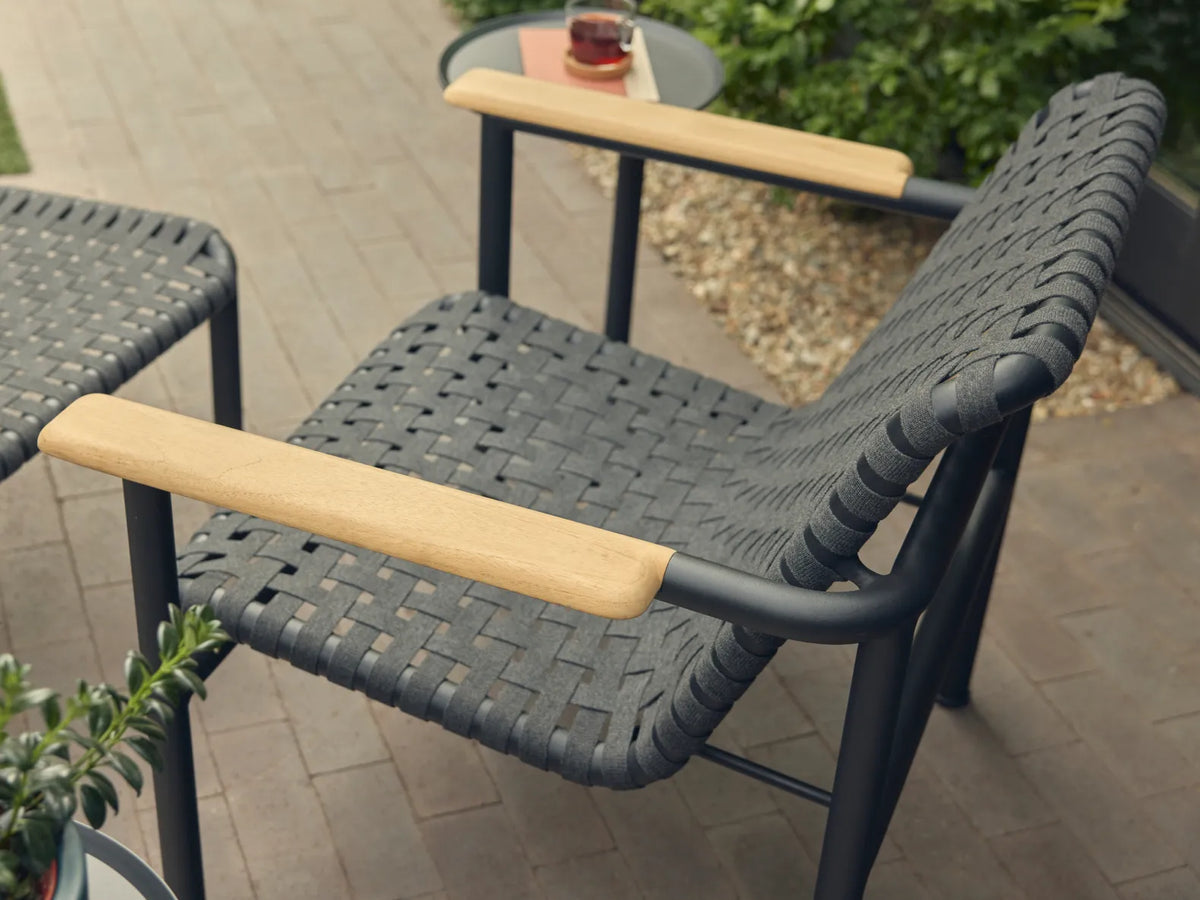 Overhead three-quarter view of a Neighbor Up-Down Chair with charcoal woven-strap seat-and-back sling and natural-wood armrests on a black powder-coated tubular frame, paired with a Neighbor Up-Down Ottoman, both on a brick-paver patio with greenery and a small black side table holding a glass and a coral-orange book in frame.