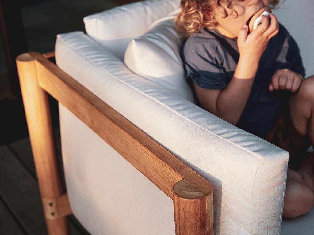 Child resting against Neighbor teak lounge chair with white cushion in a warmly lit outdoor setting