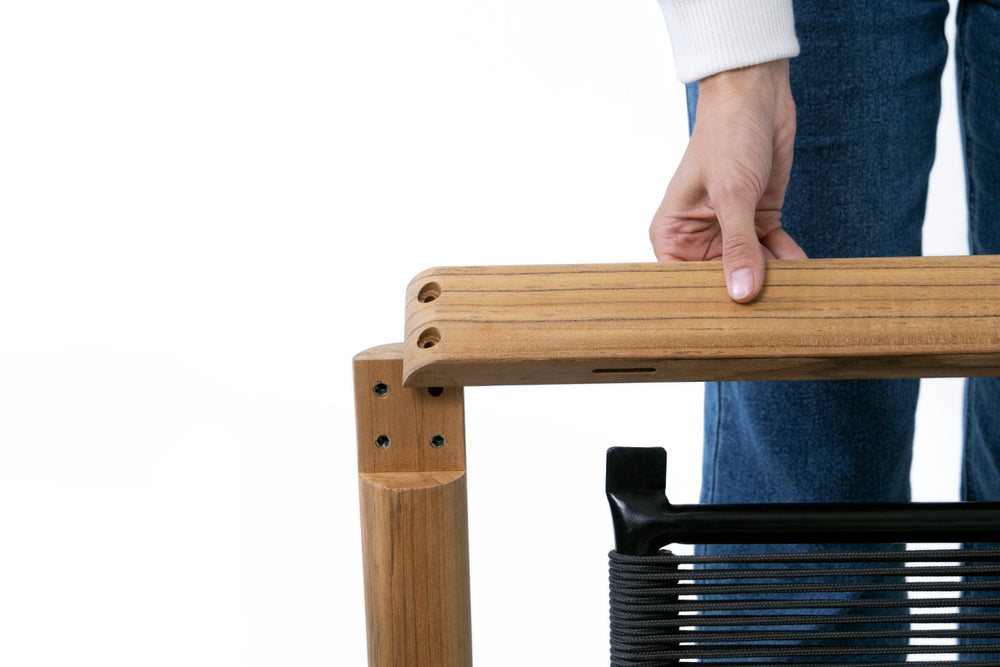 Person gripping a teak chair arm during assembly, showing teak frame joinery and dark woven cord seat