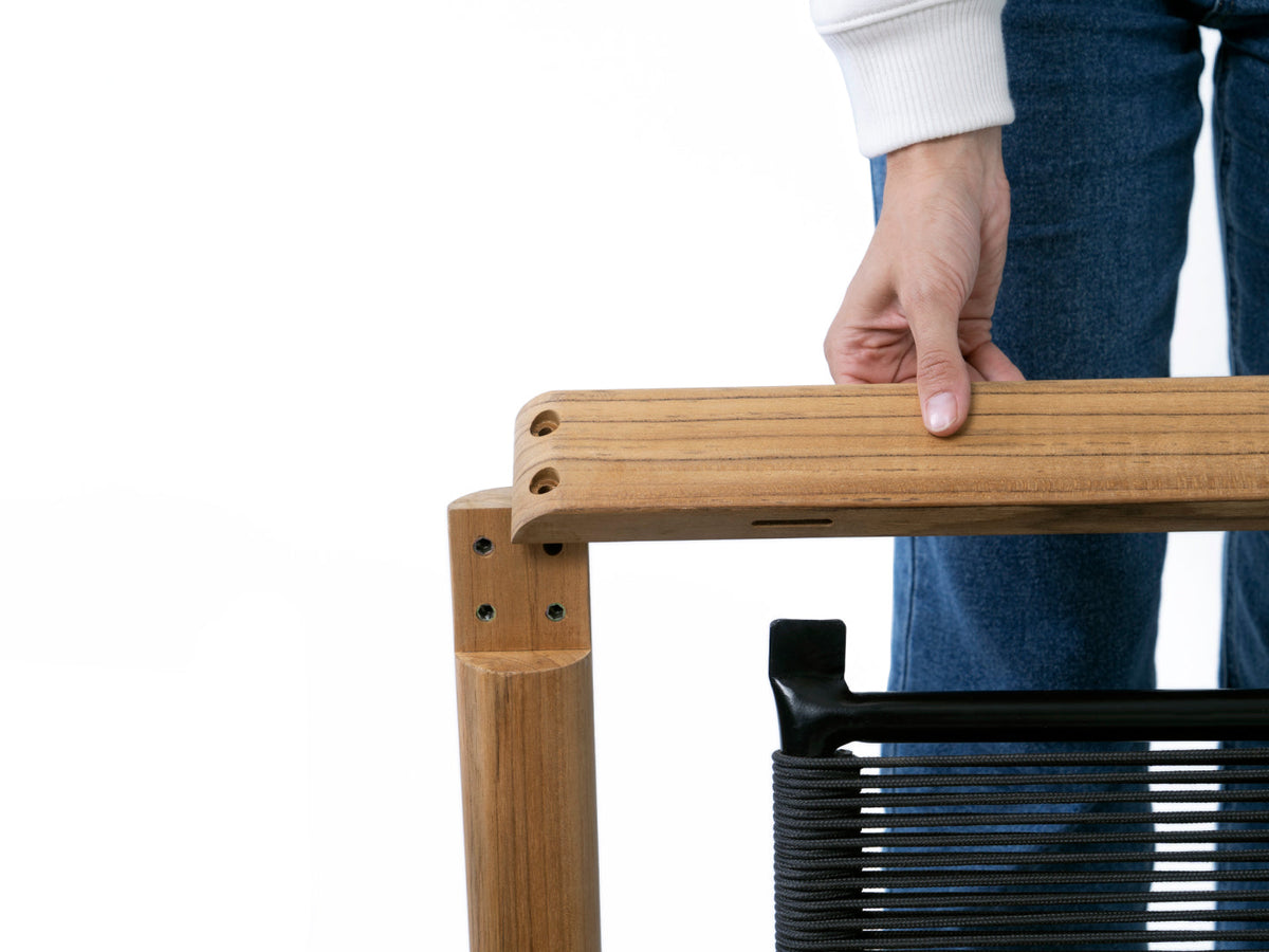 Person gripping a teak chair arm during assembly, showing teak frame joinery and dark woven cord seat
