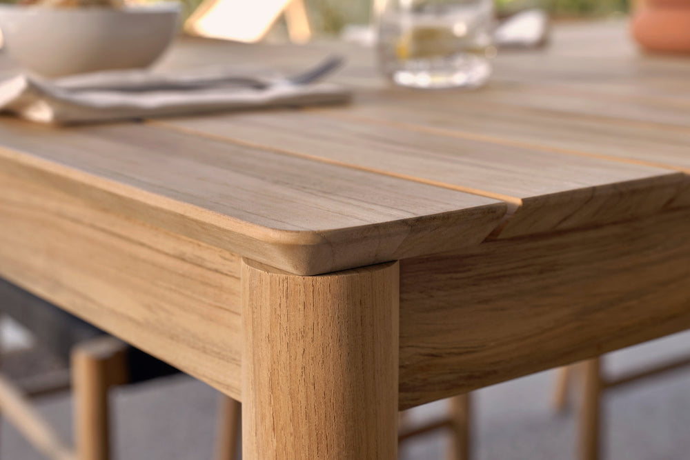Close-up corner of teak dining table surface and leg joint, place settings blurred in background