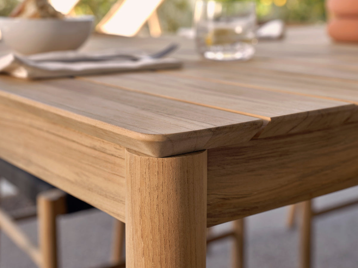 Close-up corner of teak dining table surface and leg joint, place settings blurred in background