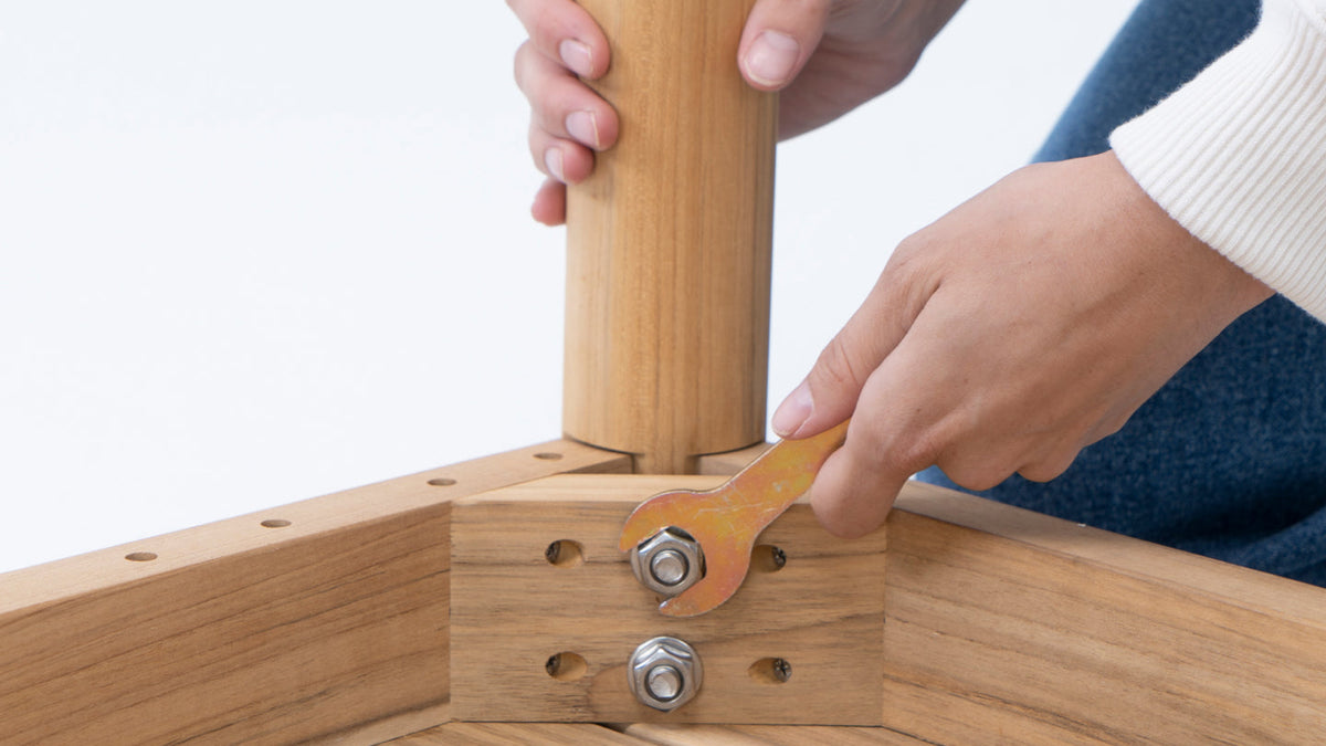 Hands assembling teak dining furniture, tightening a bolt at the table leg joint with a wrench
