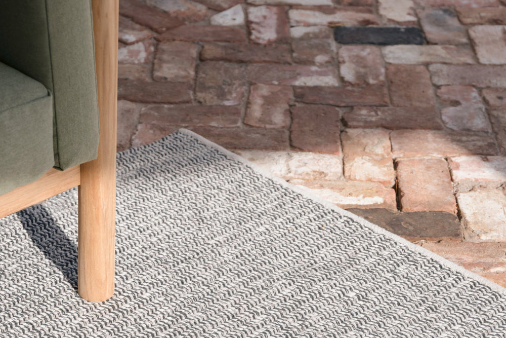 Neighbor Perf Rug in cream and grey herringbone weave on a brick paver patio, teak chair leg and green fabric chair edge visible