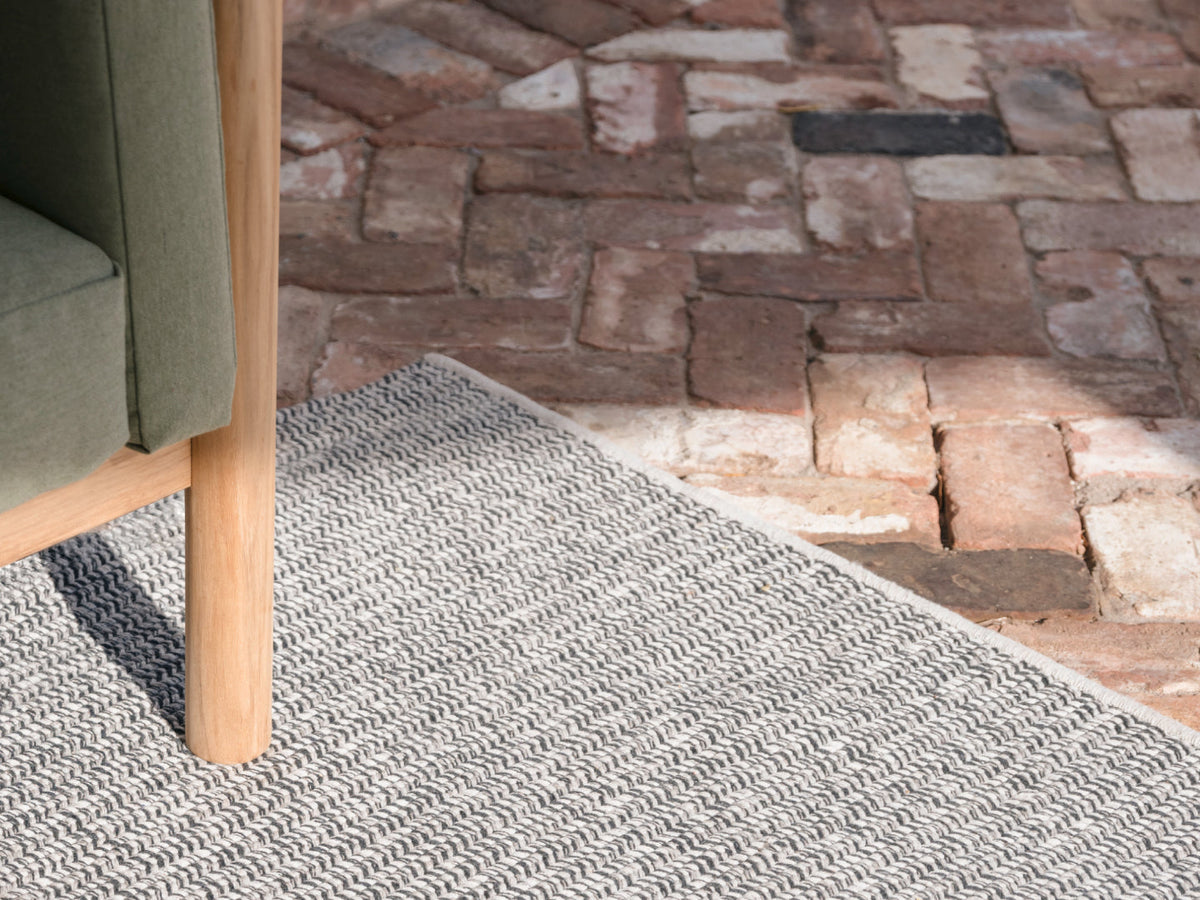 Neighbor Perf Rug in cream and grey herringbone weave on a brick paver patio, teak chair leg and green fabric chair edge visible