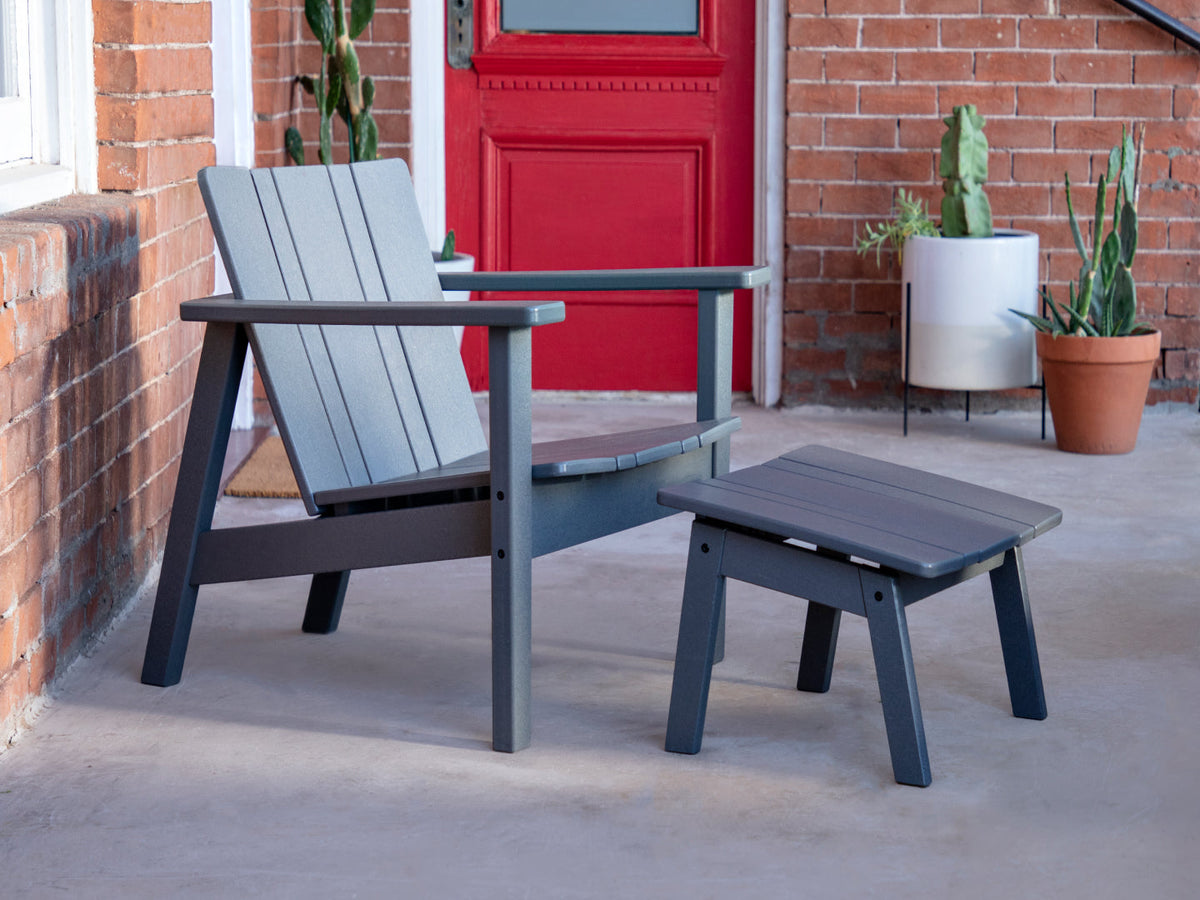 Neighbor Low Chair and Low Ottoman in charcoal polywood on a concrete front porch, red door and brick wall behind with cacti in planters