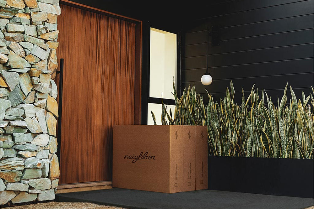 Neighbor-branded cardboard delivery box on the doorstep of a modern home with a wood door, stone cladding, and snake plant garden