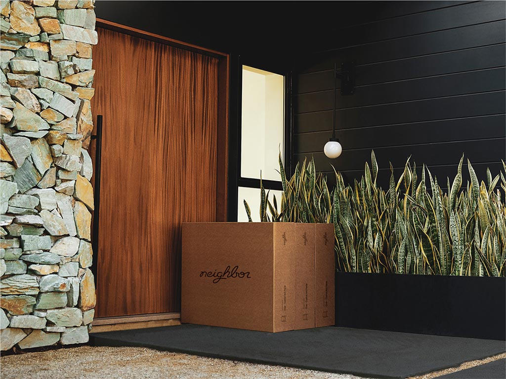Neighbor-branded cardboard delivery box on the doorstep of a modern home with a wood door, stone cladding, and snake plant garden